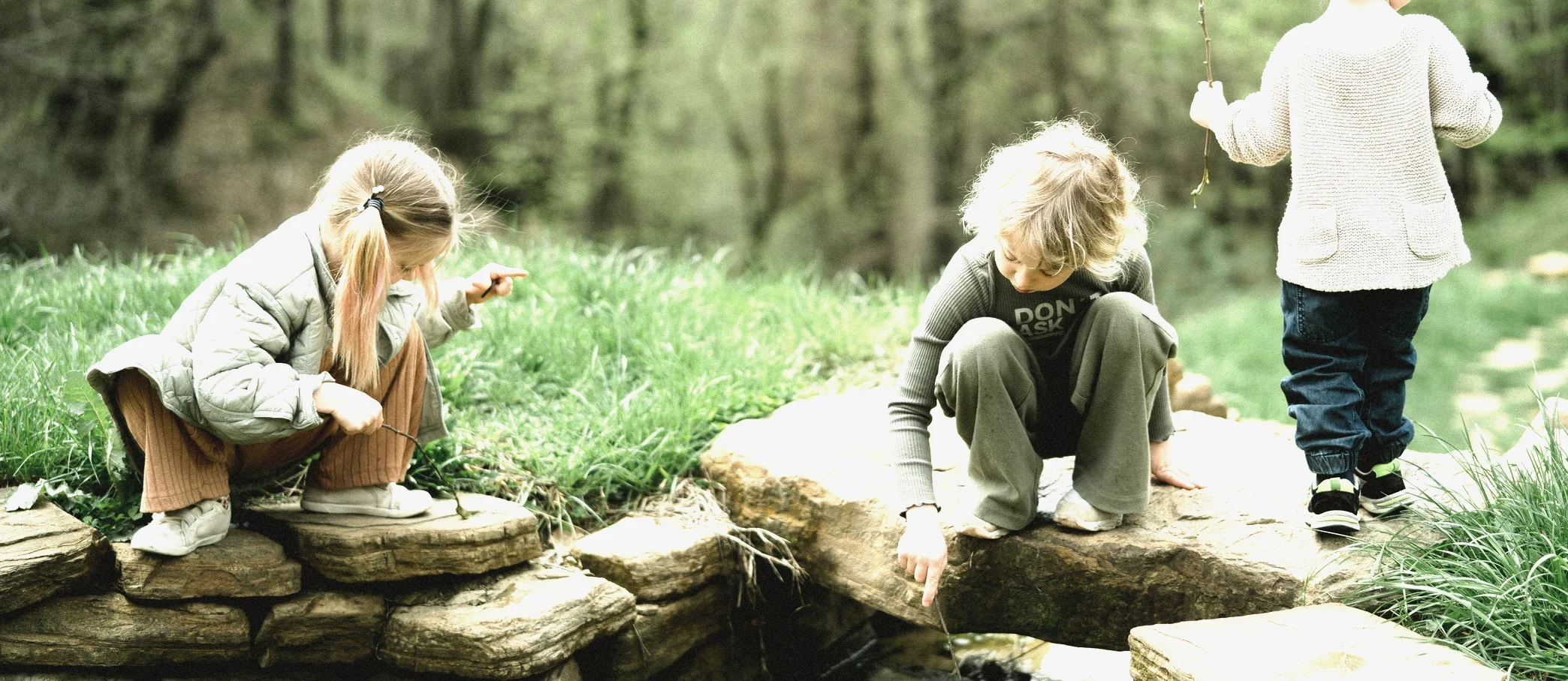 Three children crouched on rocks beside a creek, poking at the water with sticks