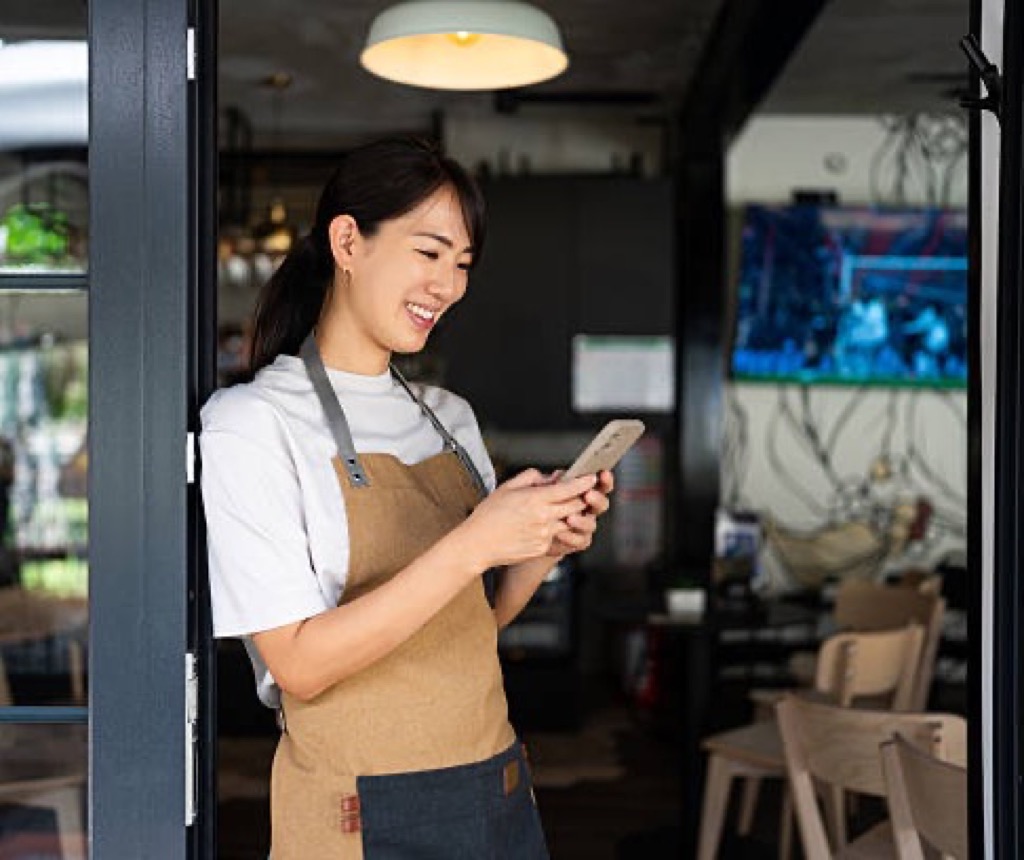 Cafe worker in an apron checking shift tasks on her phone in the doorway